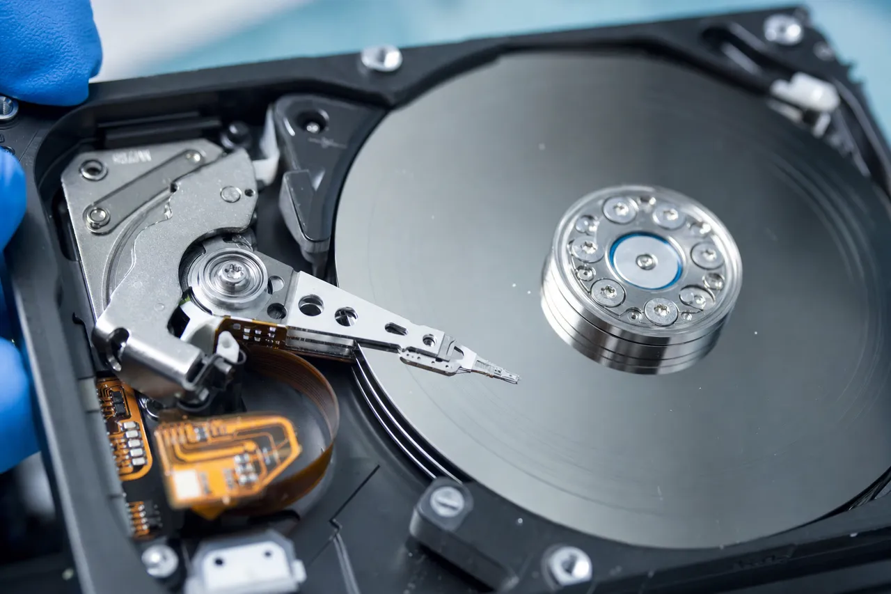 Inside an opened hard drive: the actuator arm and read/write heads parked at the edge of a mirror-finish platter showing faint concentric contact marks.