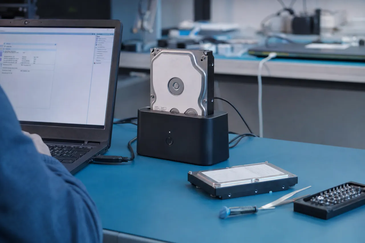 A bare hard drive in a powered SATA docking station connected to a laptop, with an empty target drive ready for cloning