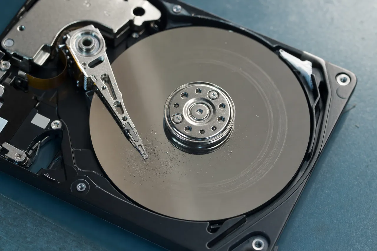 Extreme macro of an opened hard drive after a drop: the crashed read/write head slider resting on a mirror-finish platter with a faint scoring arc and fine debris near the contact point.
