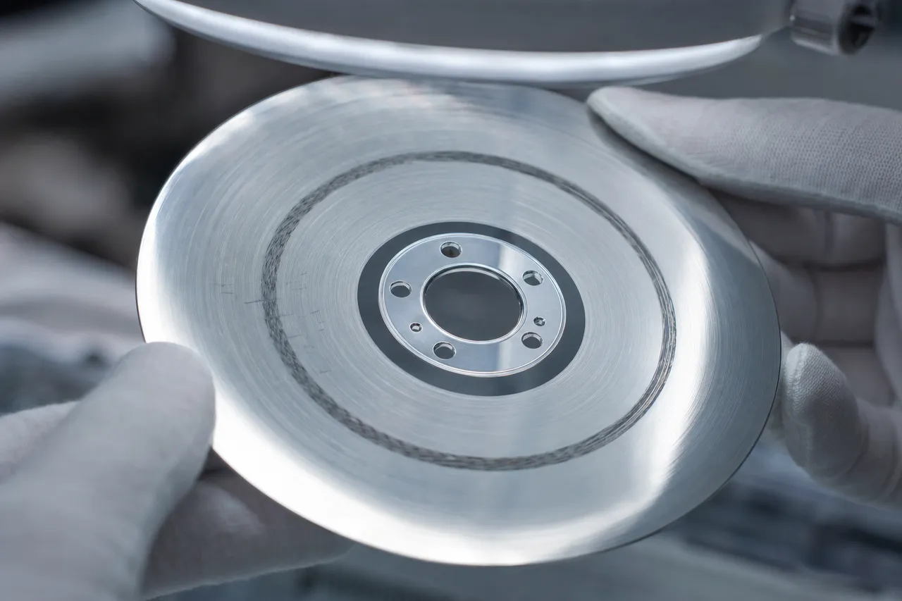 Close macro of a bare hard drive platter under a lab magnifier showing concentric scratches and a darkened scored ring where a crashed head gouged the magnetic coating.