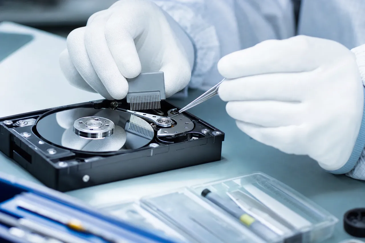 Macro of a technician using a precision head-comb tool to separate the read/write head stack from the platter surface of an opened hard drive inside a clean bench.