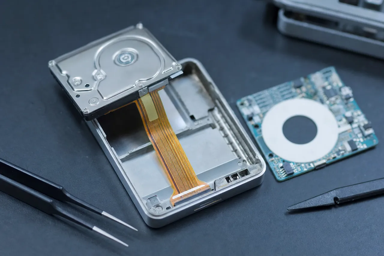 Extreme macro of a tiny 1.8-inch Toshiba hard drive lifted from an iPod Classic chassis with the delicate ZIF ribbon cable still attached, the empty aluminum shell and click-wheel board beside it.
