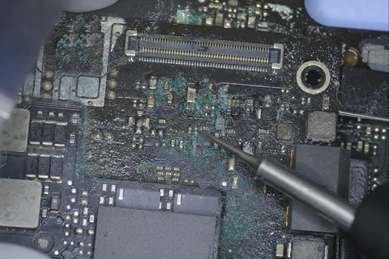 Liquid-damaged MacBook logic board with greenish corrosion spreading across the copper traces near the T2 security chip, under clinical inspection light in the MDrepairs lab before ultrasonic cleaning