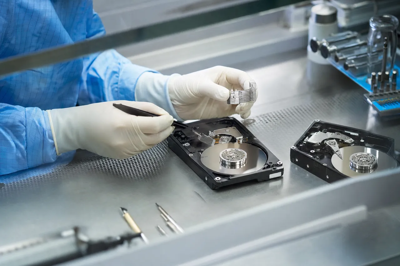 A laminar-flow clean bench in a data recovery lab with an opened legacy hard drive under bright filtered light, gloved hands using a head-comb to perform a head stack swap