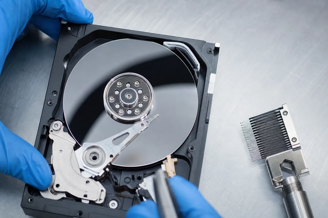 An opened legacy desktop hard drive on a clean bench showing its mirror-like aluminum platters and read-write head actuator arm with a head-comb tool resting beside it