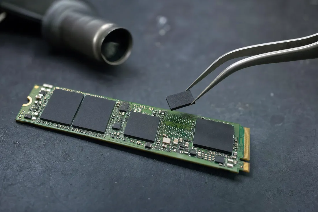 Macro view of an M.2 NVMe SSD with a single NAND flash chip lifted by tweezers above an empty solder pad for direct reading