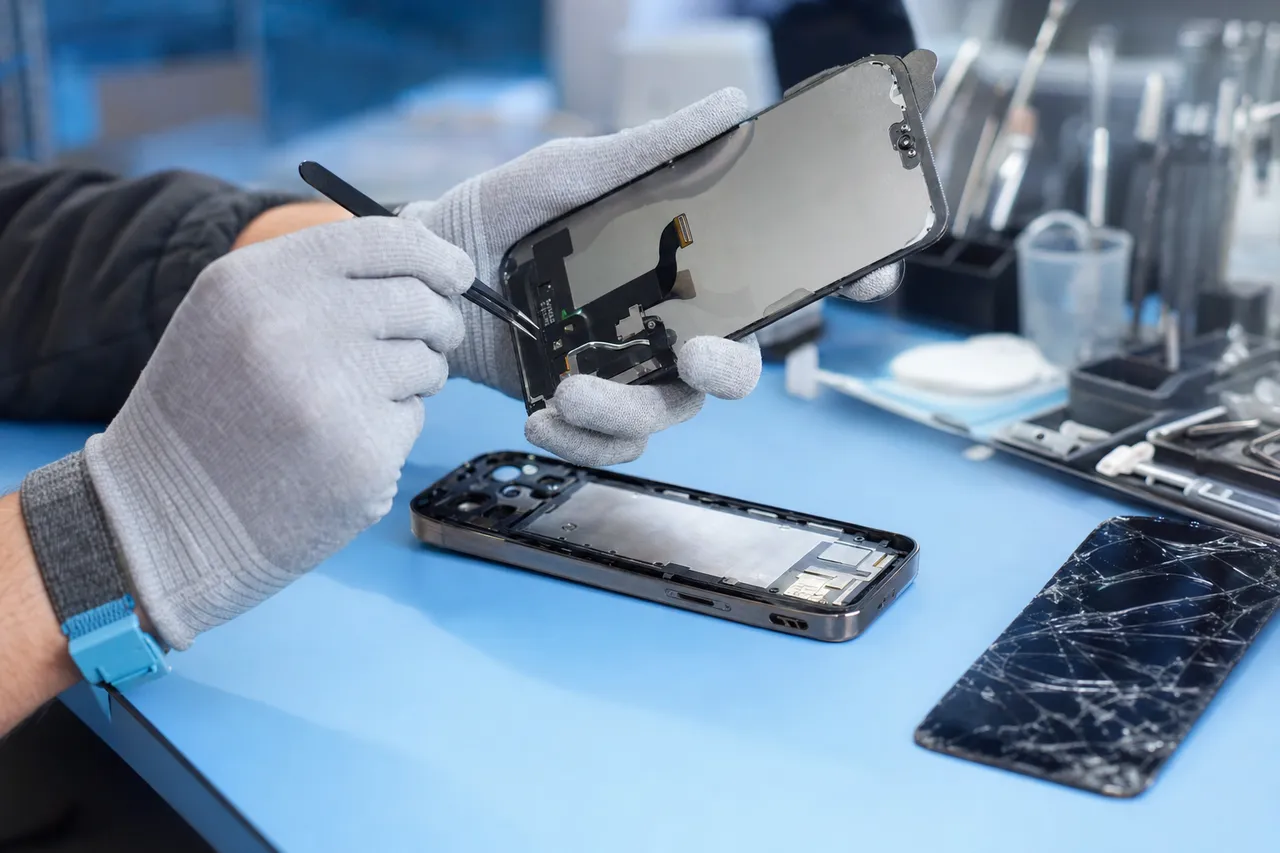 A technician lifting a replacement smartphone display assembly with its ribbon cable away from an opened phone on a repair bench