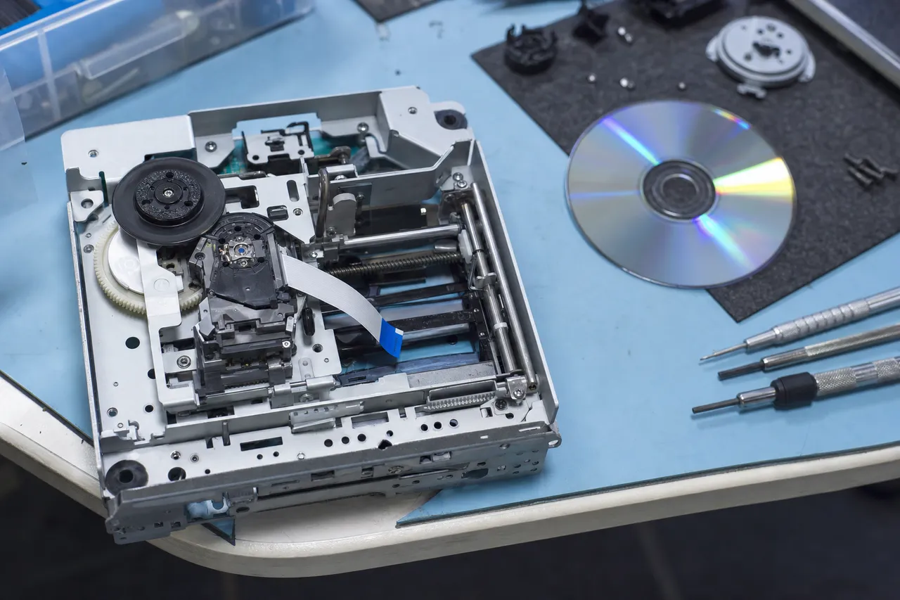 A game console optical disc drive opened on a repair bench showing the laser lens assembly, belt and gears
