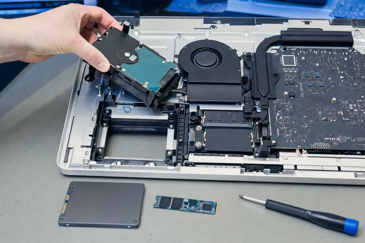 A mechanical hard drive being lifted from the interior of a thin all-in-one computer with an SSD and M.2 drive staged for an upgrade