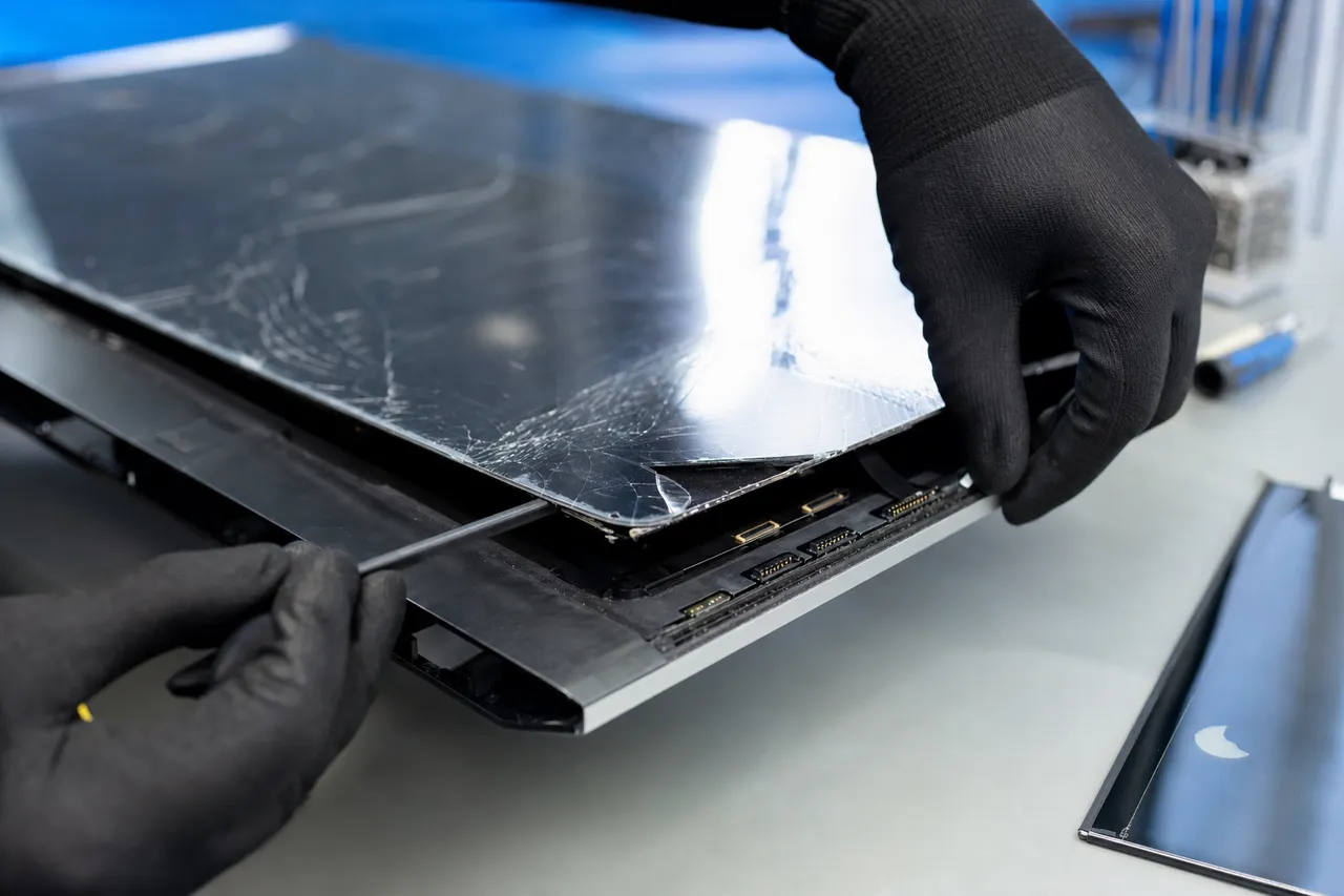 Gloved hands peeling a cracked all-in-one computer glass and LCD display assembly away from its aluminum body on a repair bench