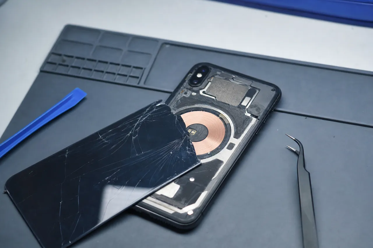 An iPhone laid face-down with its cracked rear glass partially removed exposing the wireless charging coil and internal frame