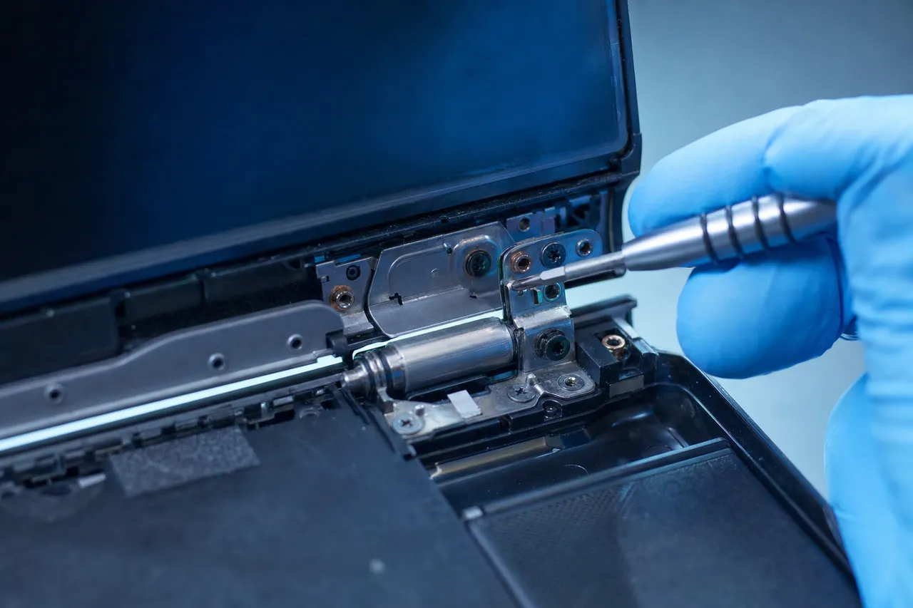 Extreme close-up of a laptop hinge mechanism with the bezel removed, a gloved hand tightening the metal hinge bracket with a precision screwdriver
