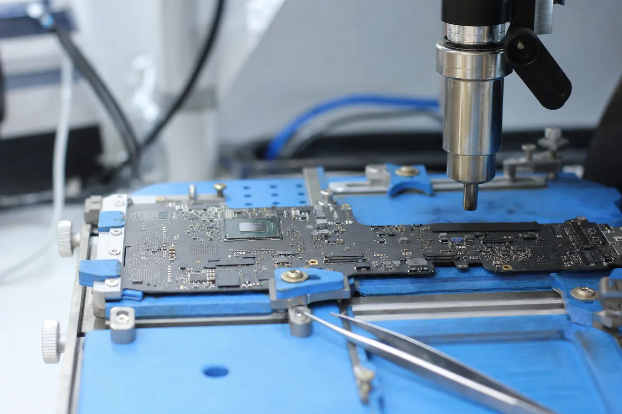 A thin MacBook logic board clamped in a board-holder fixture on a clean repair bench with precision tools nearby