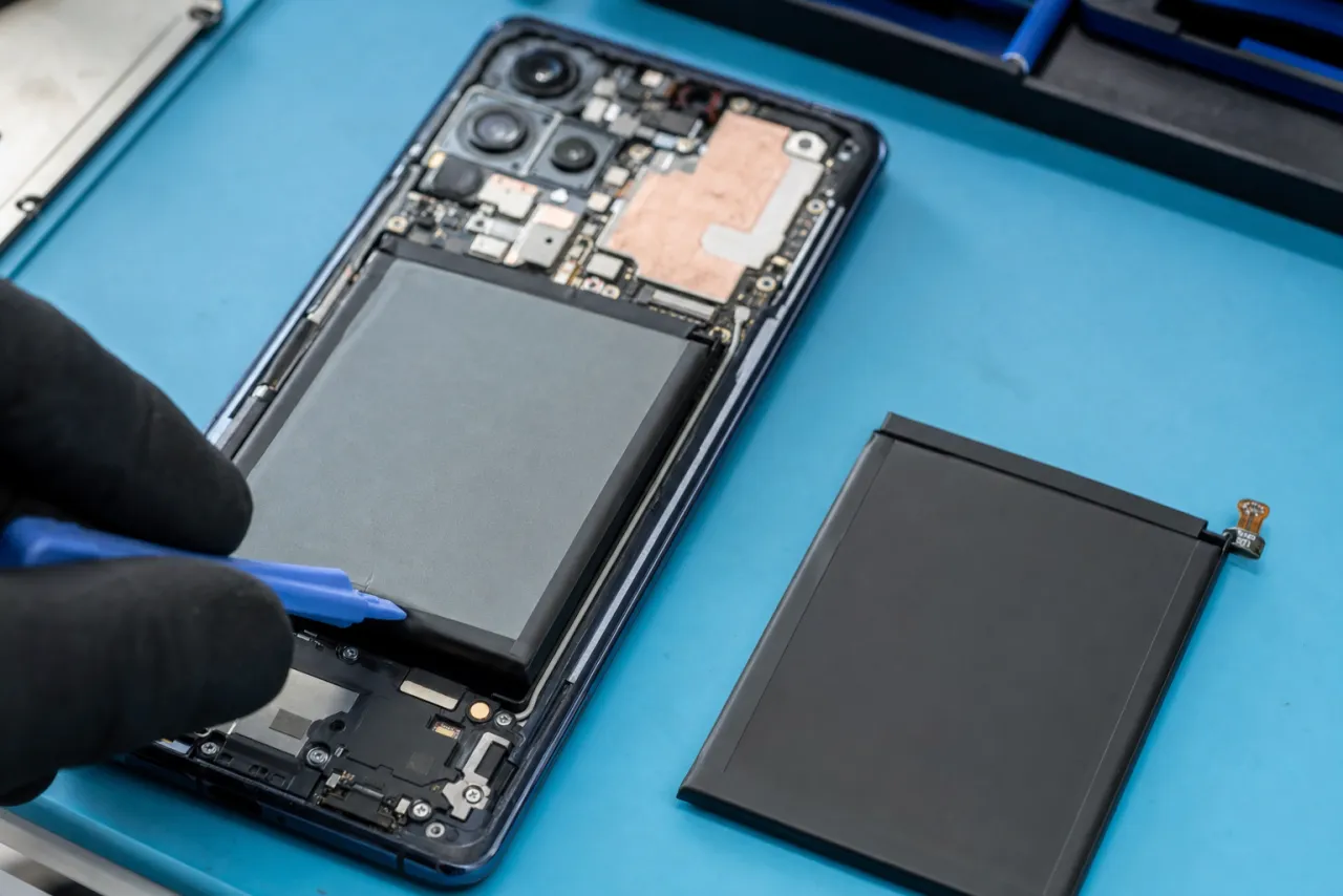An opened Galaxy phone on a repair bench with the rear glass removed exposing the battery and logic board, a pry tool lifting the battery