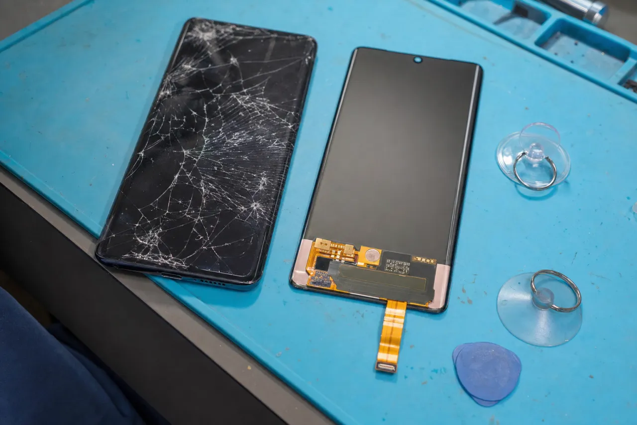 A slim Galaxy flagship with a curved-edge AMOLED display and a shattered screen on a repair bench beside a replacement display assembly