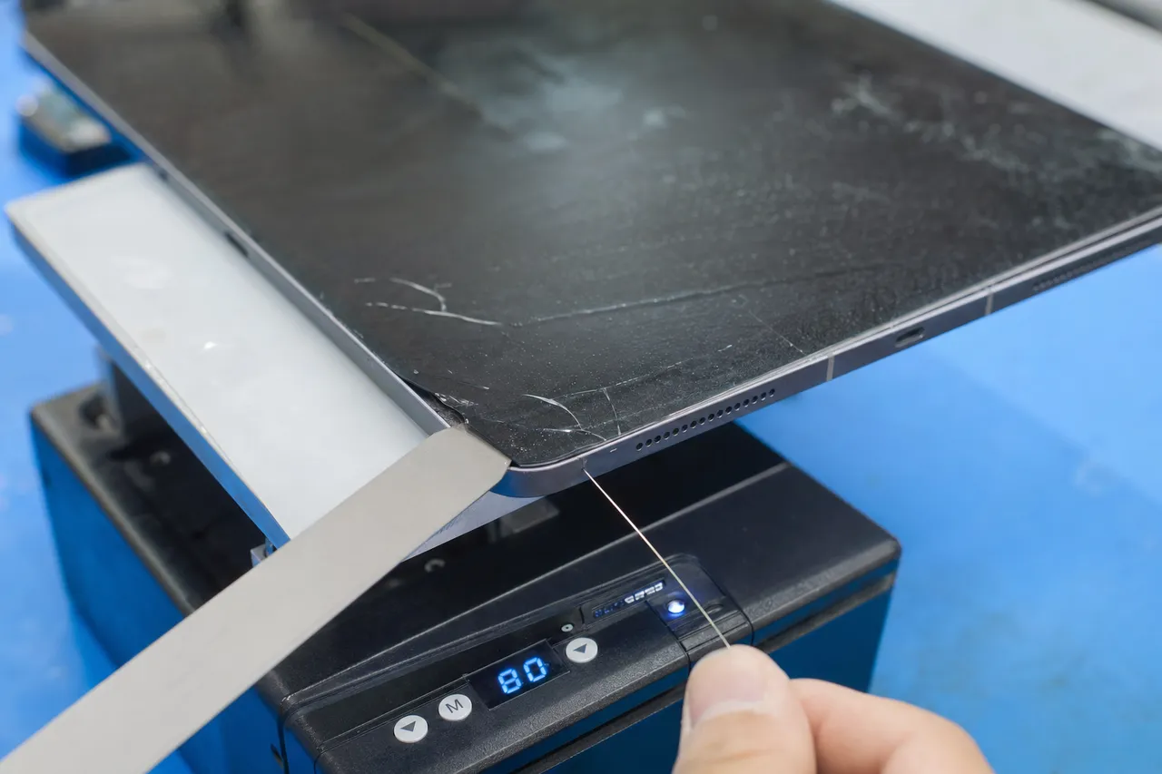A tablet face-down on a heat plate while a thin pry blade separates the cracked outer glass digitizer from the LCD panel beneath