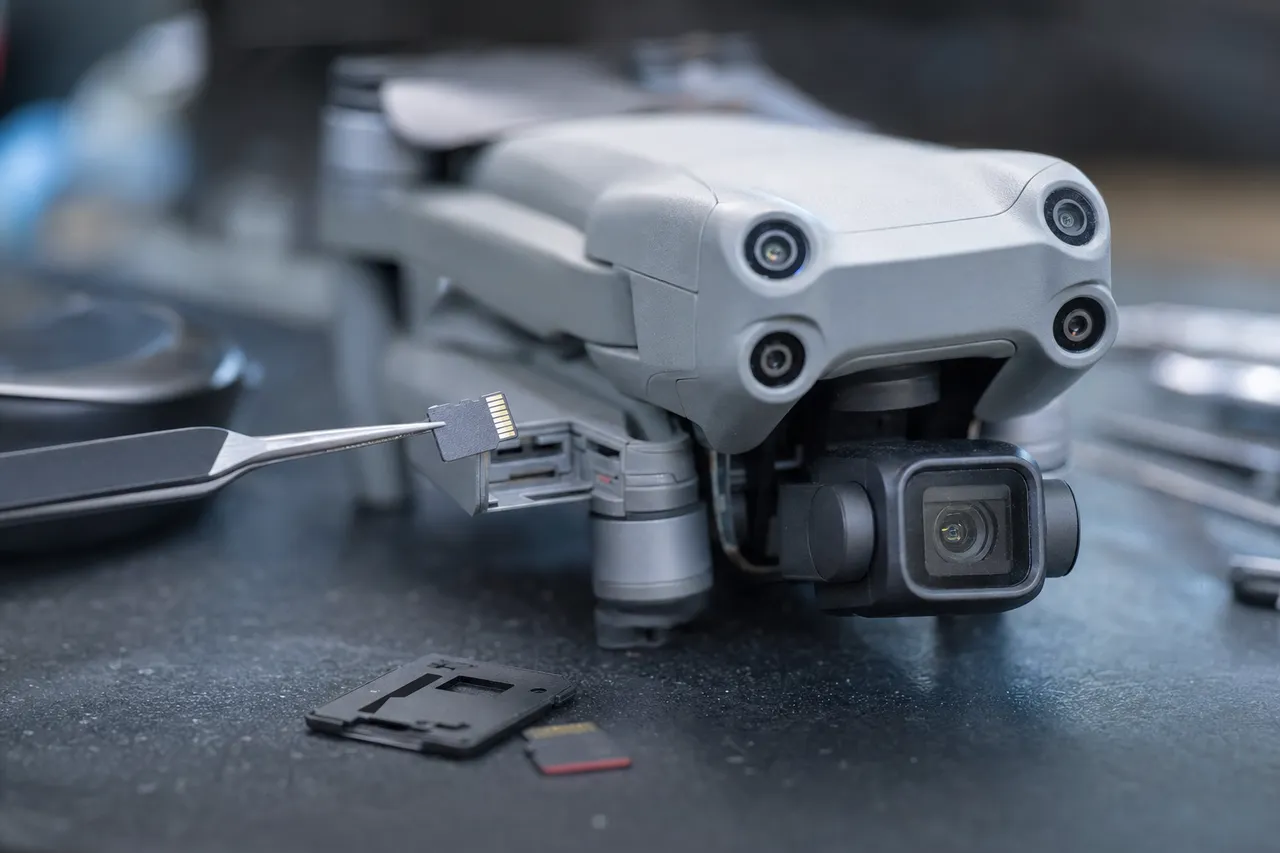 A folded camera drone with its microSD card lifted from the slot by tweezers on a lab bench