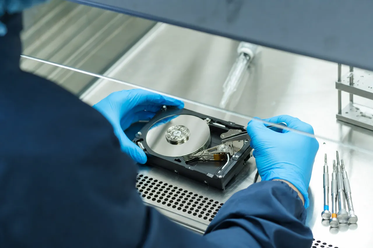A technician in blue nitrile gloves working on an opened hard drive under an ISO Class 5 laminar-flow hood, the platter stack and head actuator visible
