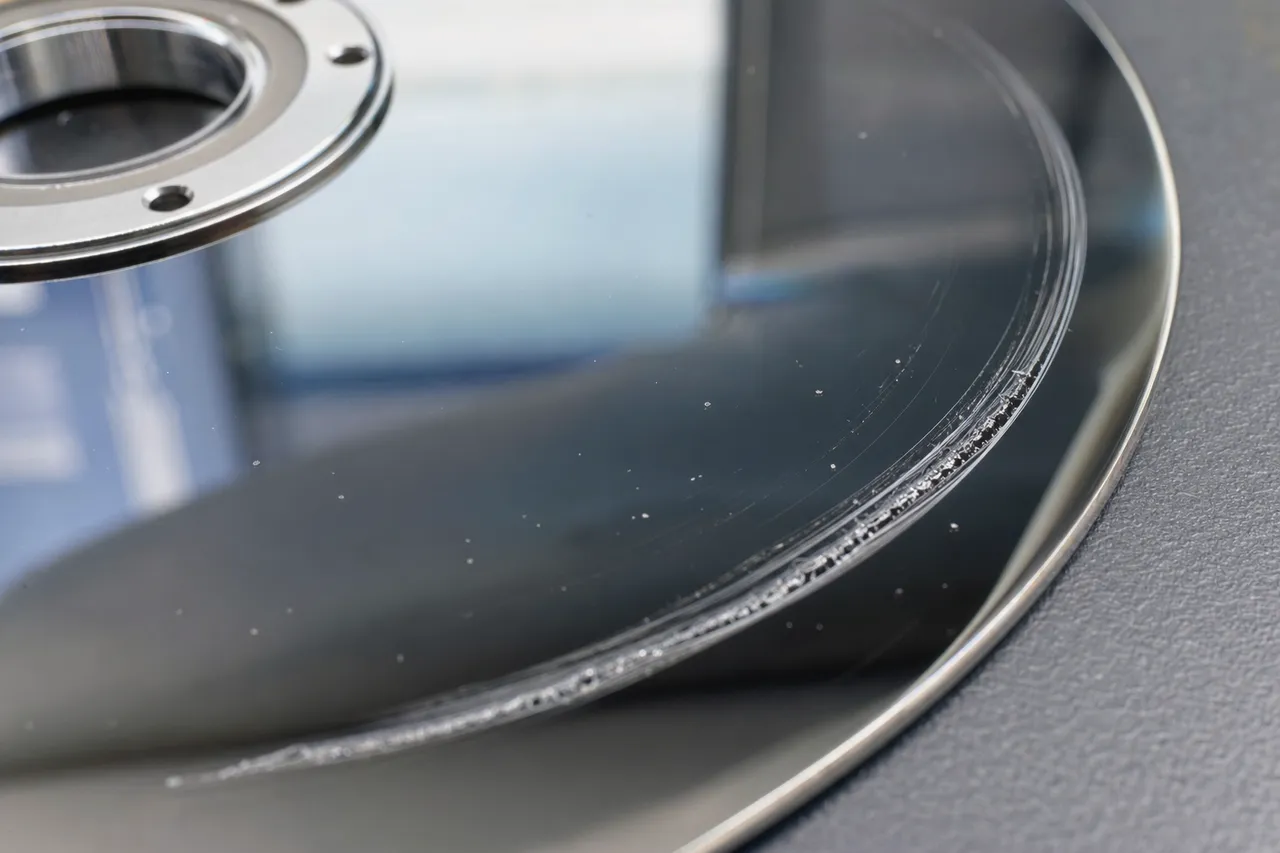 Macro close-up of a mirror-finish hard drive platter with a deep concentric head-crash scratch catching the light, fine metallic debris along the gouge
