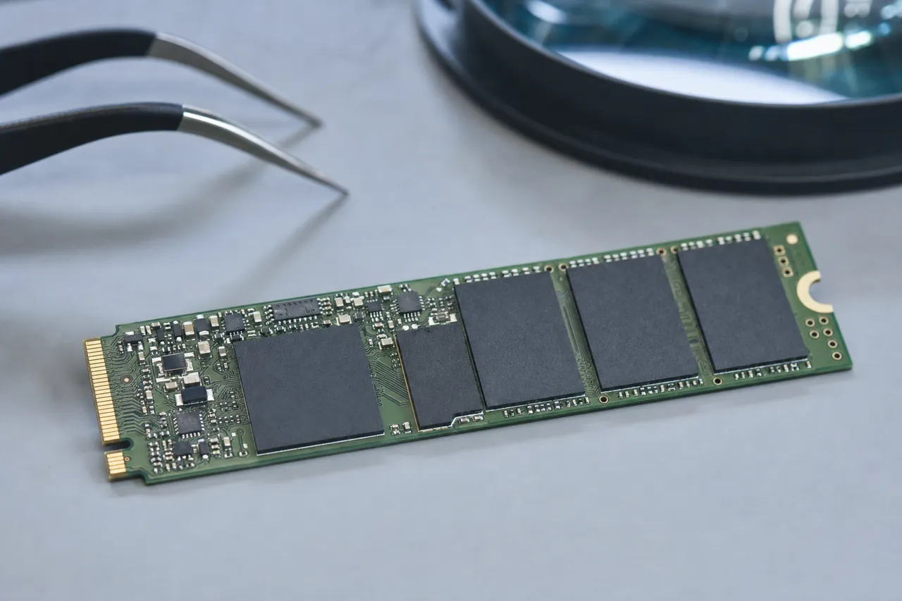 A bare M.2 NVMe solid state drive with its controller and NAND flash packages exposed, beside fine tweezers and a magnifier during a multi-stage board diagnosis